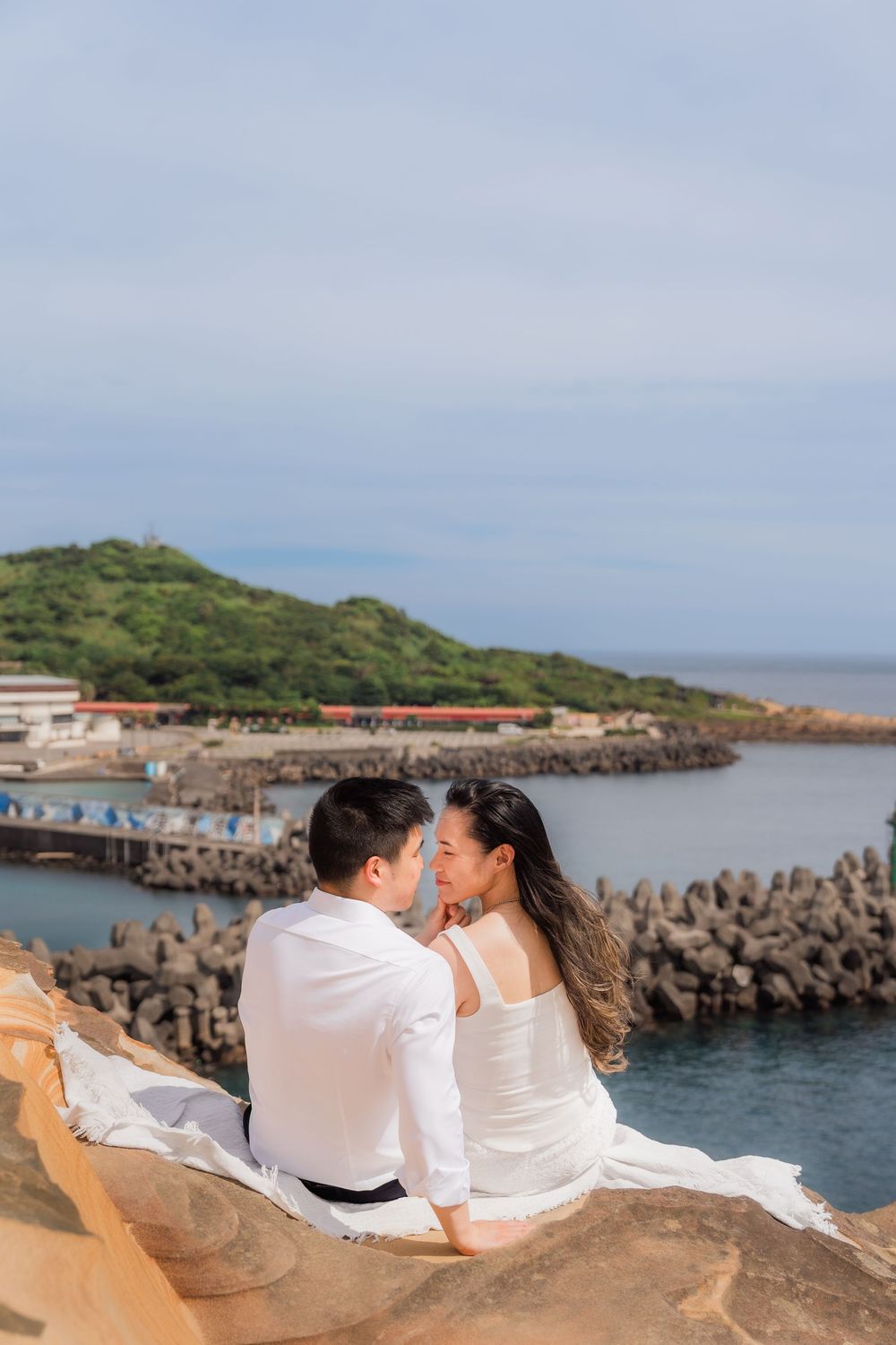 Couple in white sitting on rocks overlooking a scenic harbor with green hills and calm blue waters.