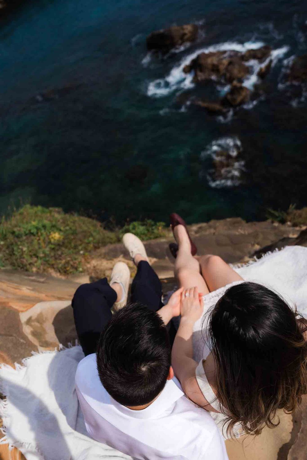 Aerial view of couple sitting on blanket at cliff edge overlooking turquoise ocean waters.