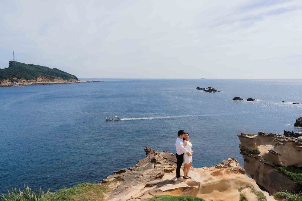 Couple embracing on rocky coastal cliffs overlooking a serene blue ocean with green hills and boats in the background.