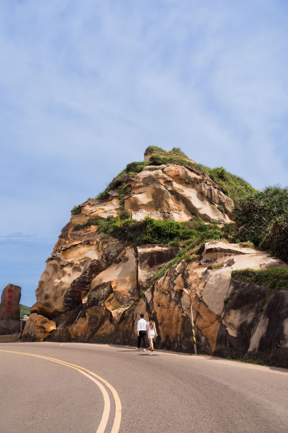 Couple walking along a coastal road beside a large rocky outcrop with green vegetation under a blue sky.