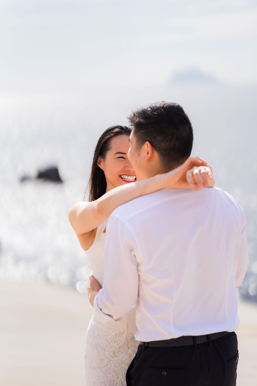 A happy couple embraces and laughs together on a sunny beach, the woman in a white dress with her arms around the man.