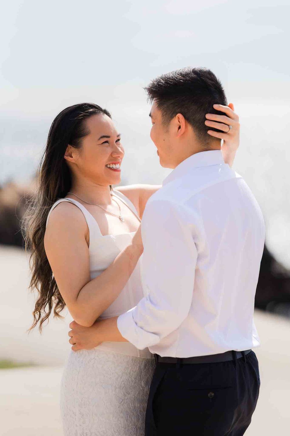 A smiling couple embraces outdoors near the ocean, woman in white dress, man in white shirt and dark pants.