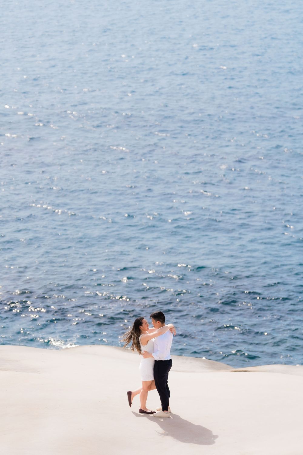 Romantic couple embracing on white sand dunes with sparkling blue ocean water in the background.