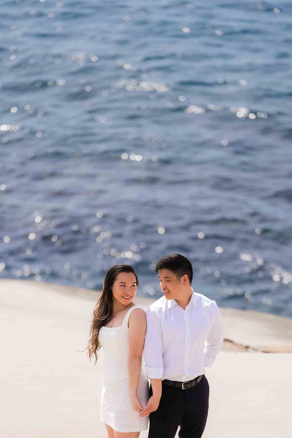 A couple in white outfits holds hands on a sunny beach with sparkling blue ocean water in the background.