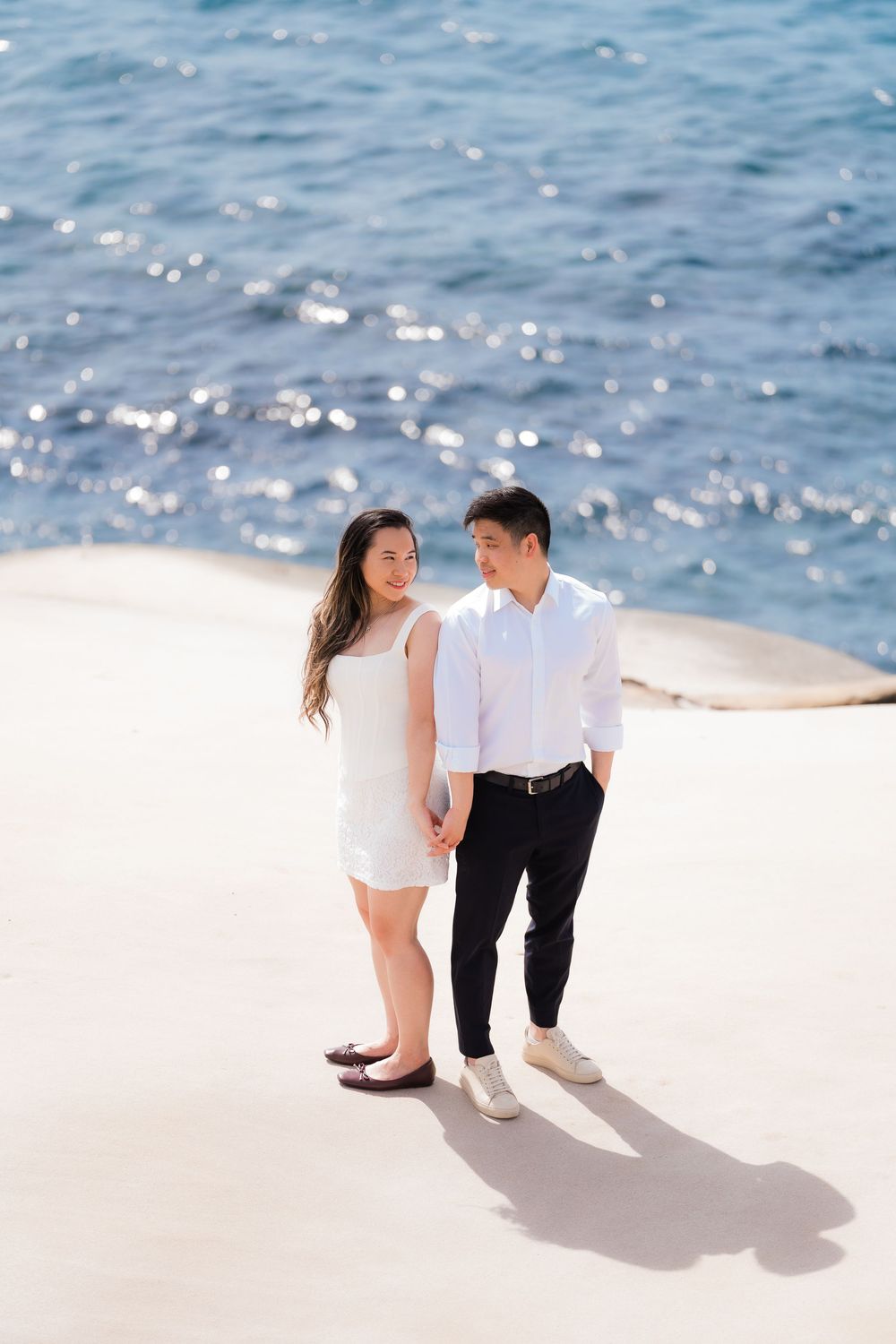 Couple holding hands on sandy shore with sparkling blue ocean backdrop, woman in white dress and man in light pink shirt.