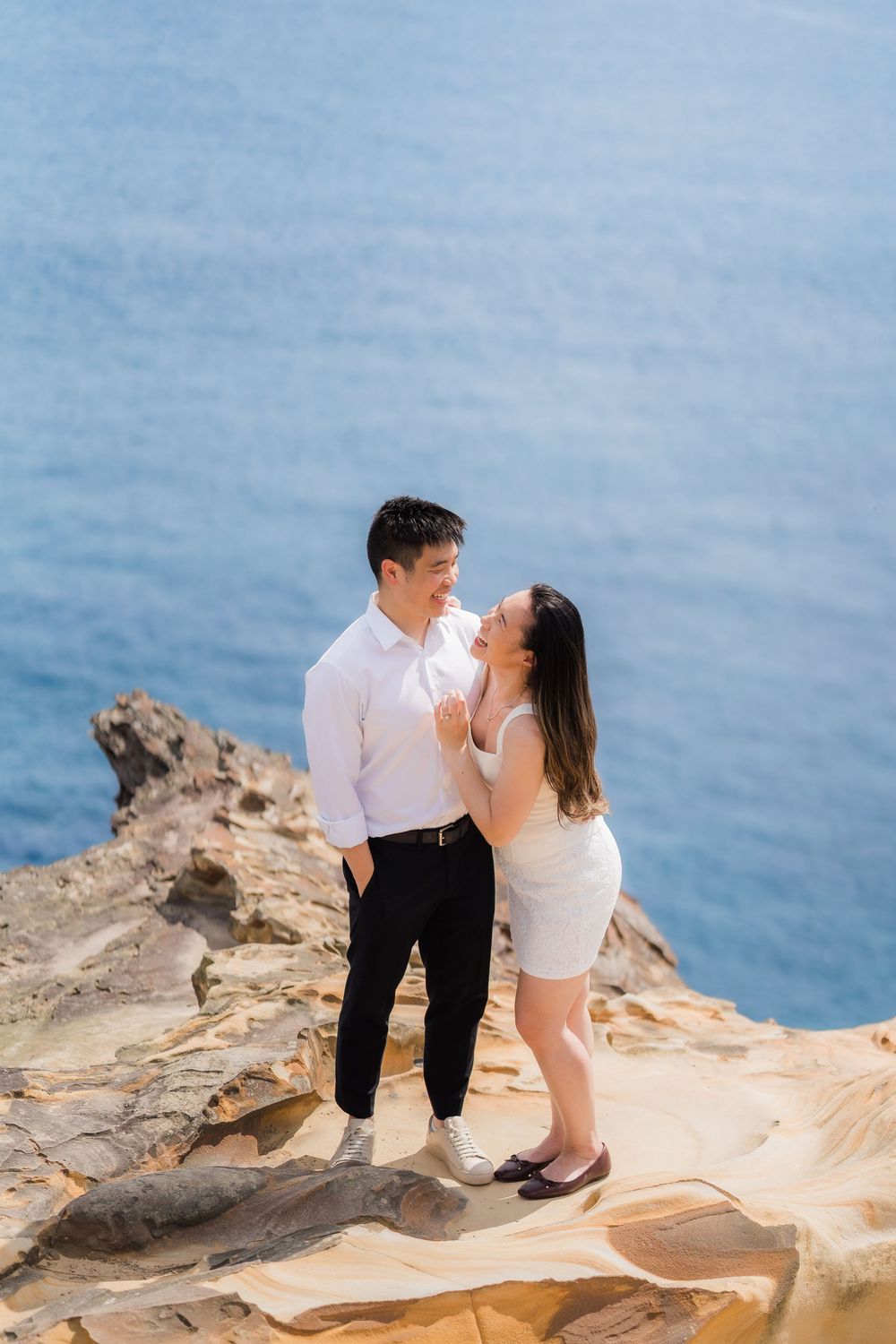 Couple in white outfits standing on rocky coastal cliff with blue ocean backdrop.