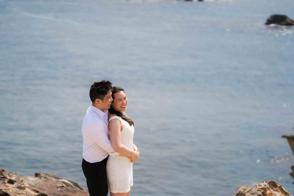 Couple embracing romantically on rocky coastal cliffs with blue ocean waves in the background.