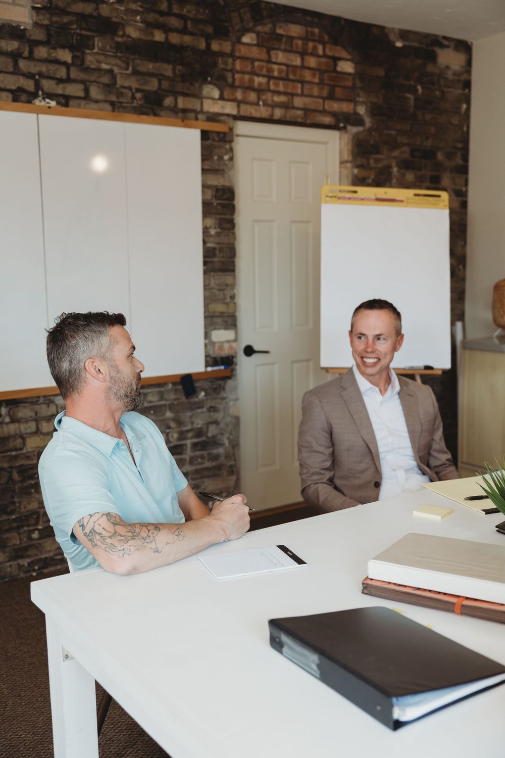 Two men smiling and conversing at a white desk in a modern office with exposed brick walls and whiteboards.