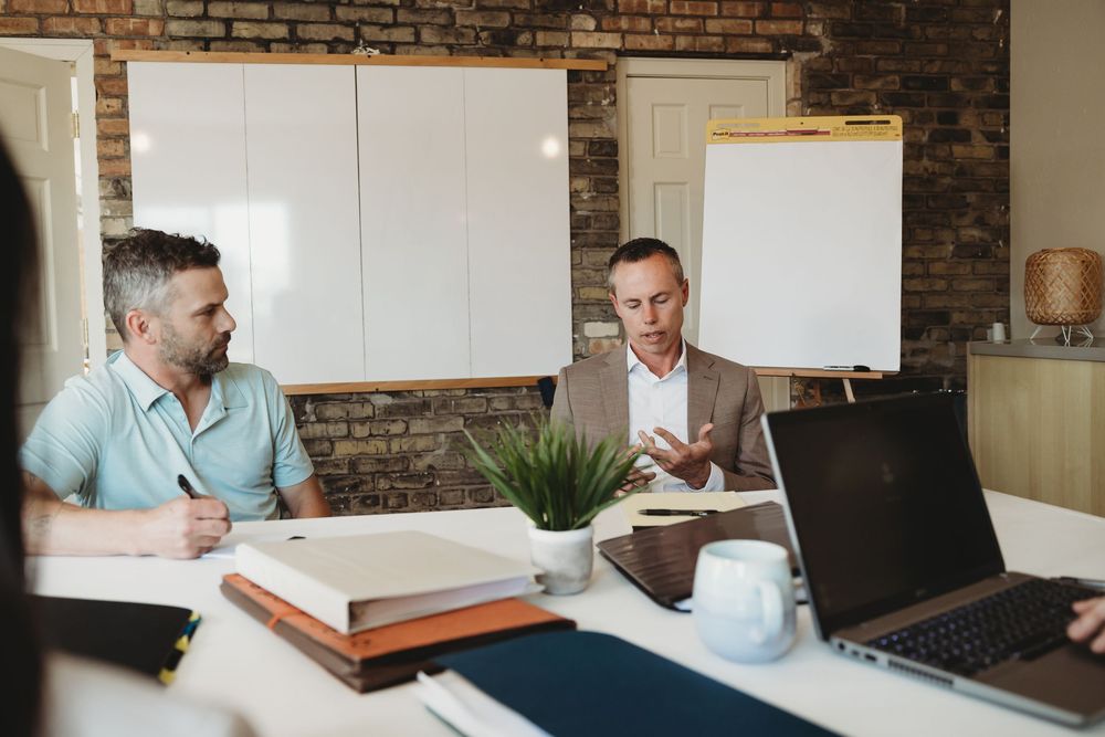 Two businessmen in a meeting room discussing strategy with laptops and notebooks on a white table.