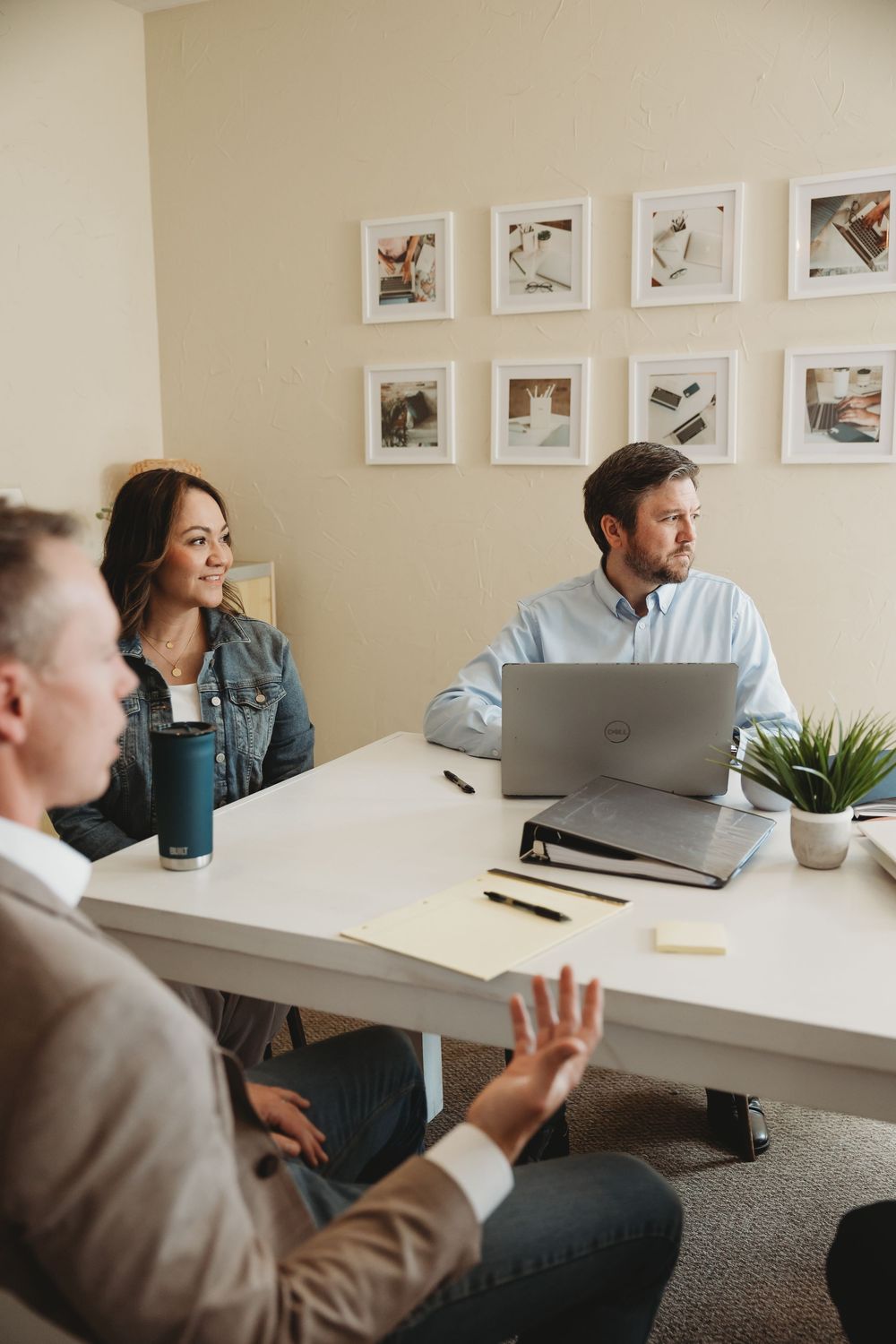 Three professionals collaborate in a bright office meeting room with a laptop, notebooks, and framed photos on the wall.