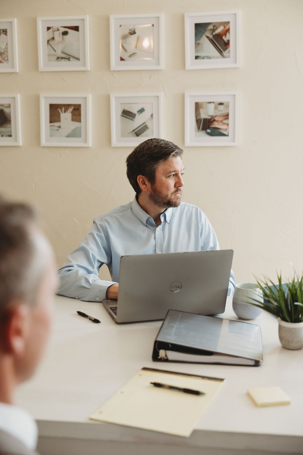 Man in light blue shirt working on laptop at desk with binder and plant, framed photos on wall behind him.