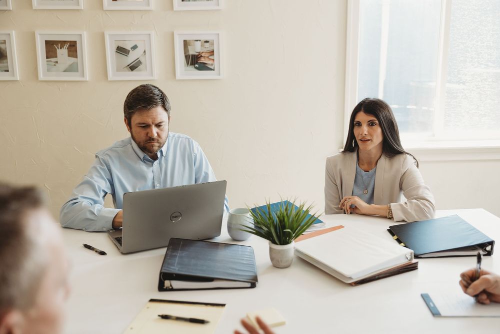 Business professionals in a meeting room, man using laptop, woman listening attentively at a white conference table.