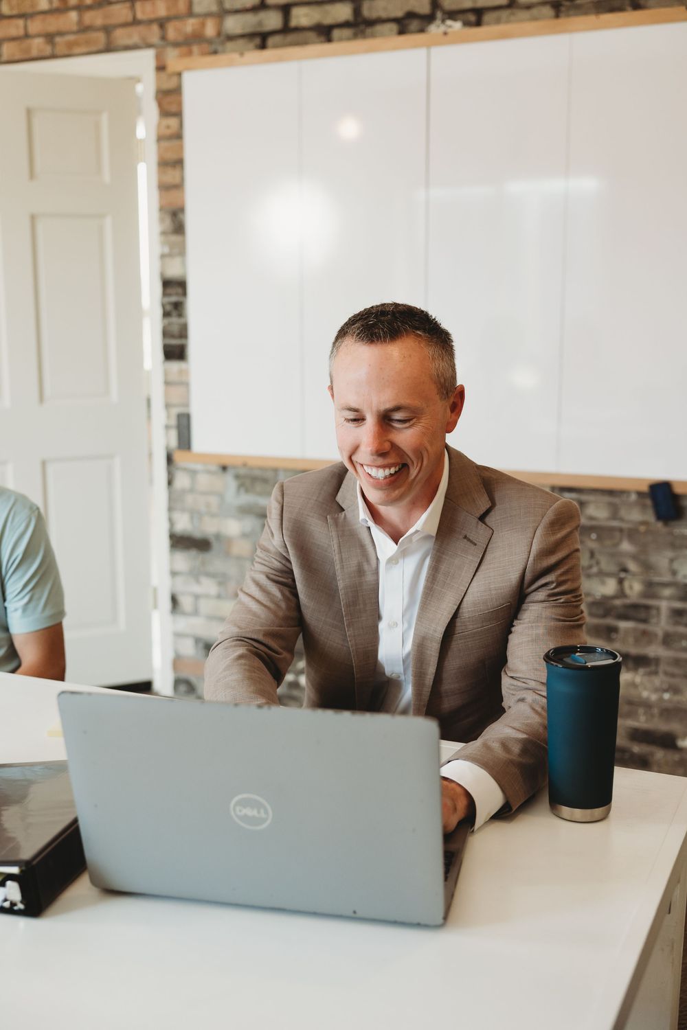 Smiling businessman in beige suit working on Dell laptop at white desk with coffee tumbler in modern brick office.