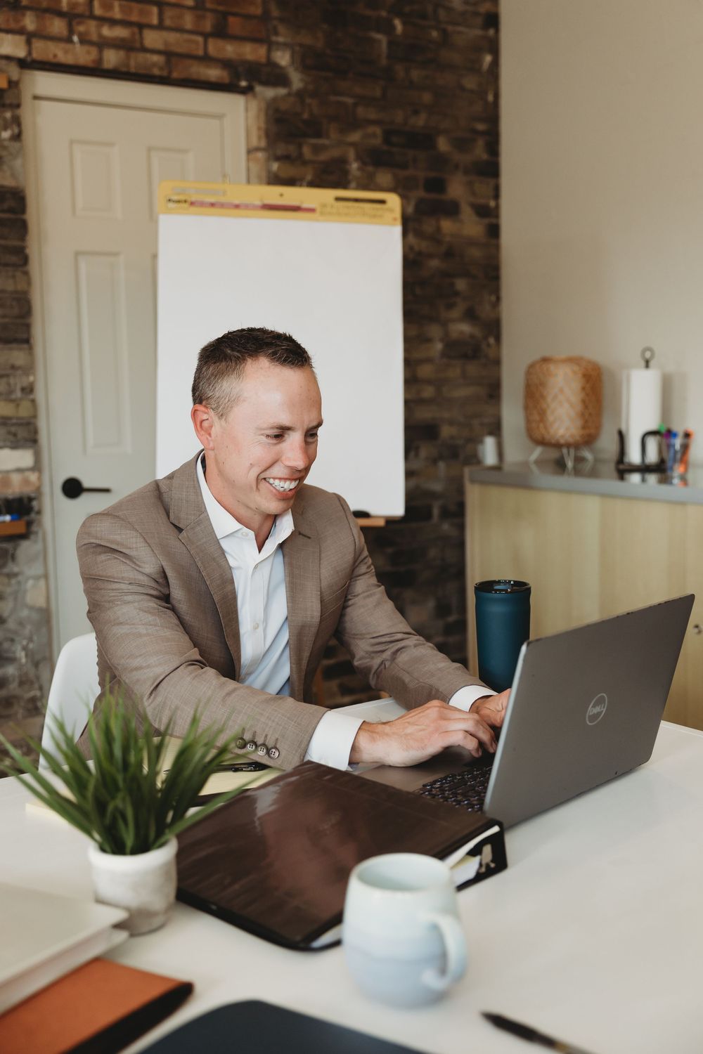Smiling businessman in tan suit working on Dell laptop at white desk with coffee mug and plant in modern office.