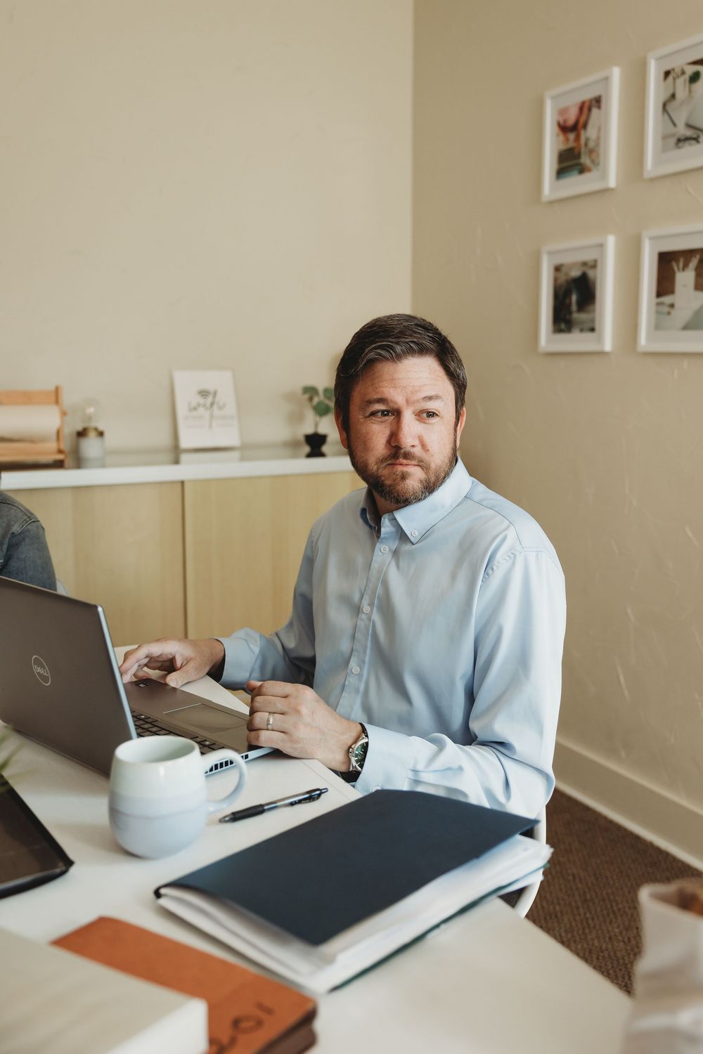 Professional man in blue shirt working on Dell laptop at desk with coffee mug and notebook in home office.