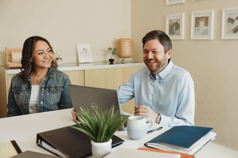 Two colleagues smiling and laughing together at a desk with a laptop, coffee, and plant in a bright office.