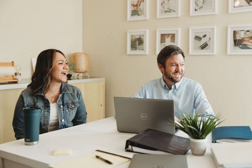 Two colleagues laughing and smiling while working together on a laptop at a modern home office desk.