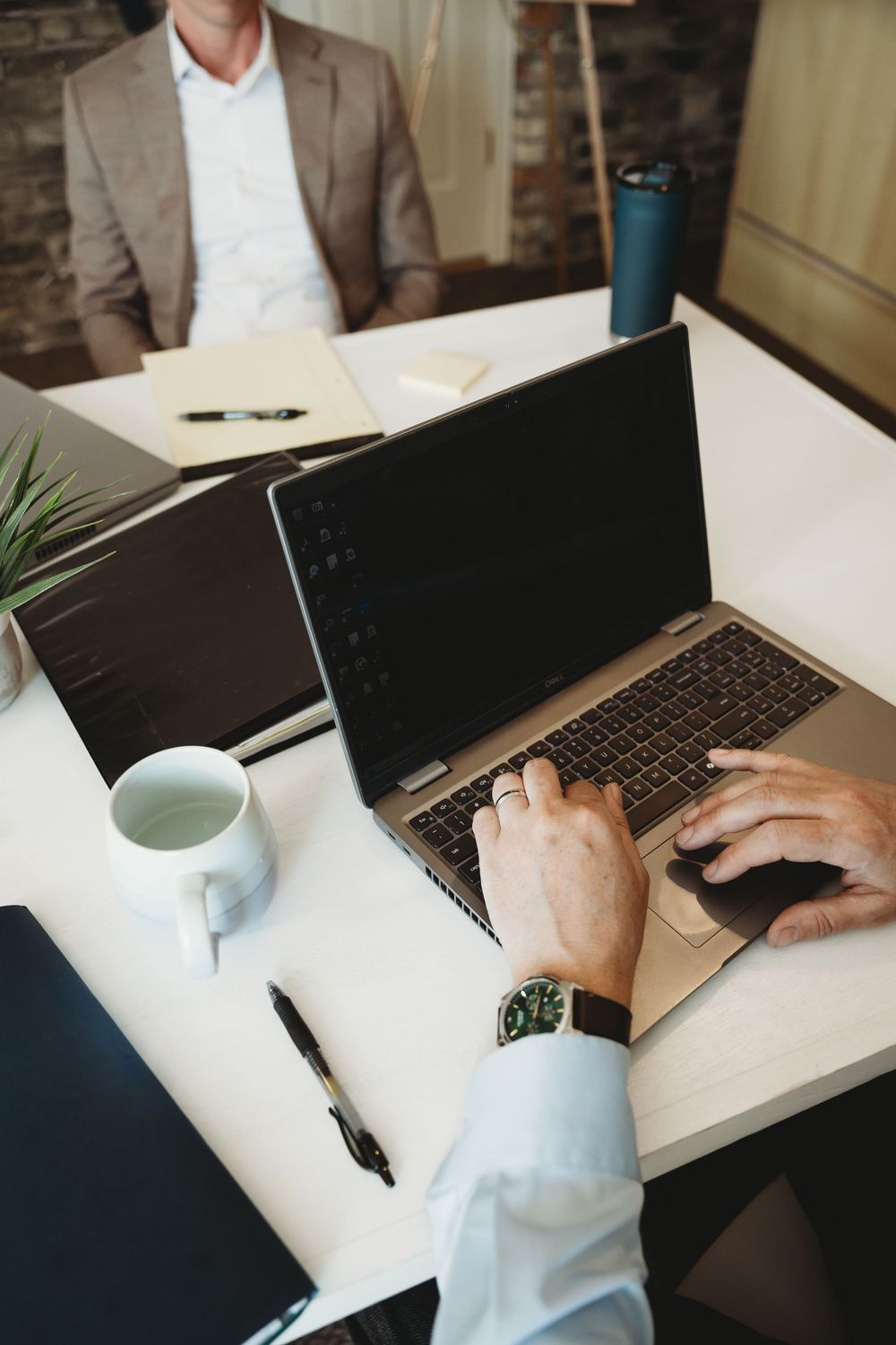 Businessman typing on laptop at office desk with coffee cup, pen, and second laptop during a business meeting.