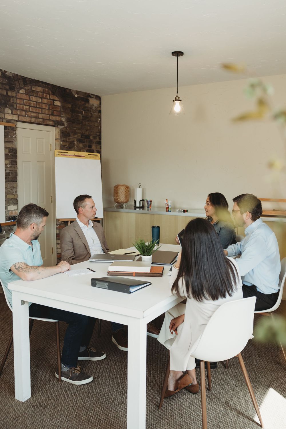 Six professionals engaged in a business meeting around a white table in a modern office with exposed brick walls.