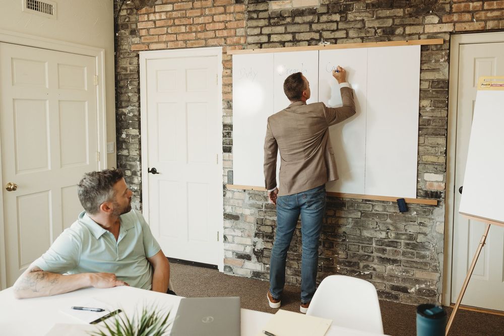 Businessman writing on a whiteboard during a meeting while a colleague watches in a modern brick-walled office.