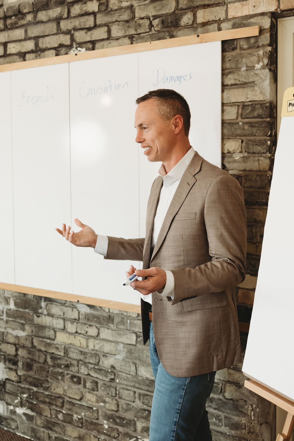 A smiling man in a blazer presents at a whiteboard in a modern brick-walled office during a business meeting.