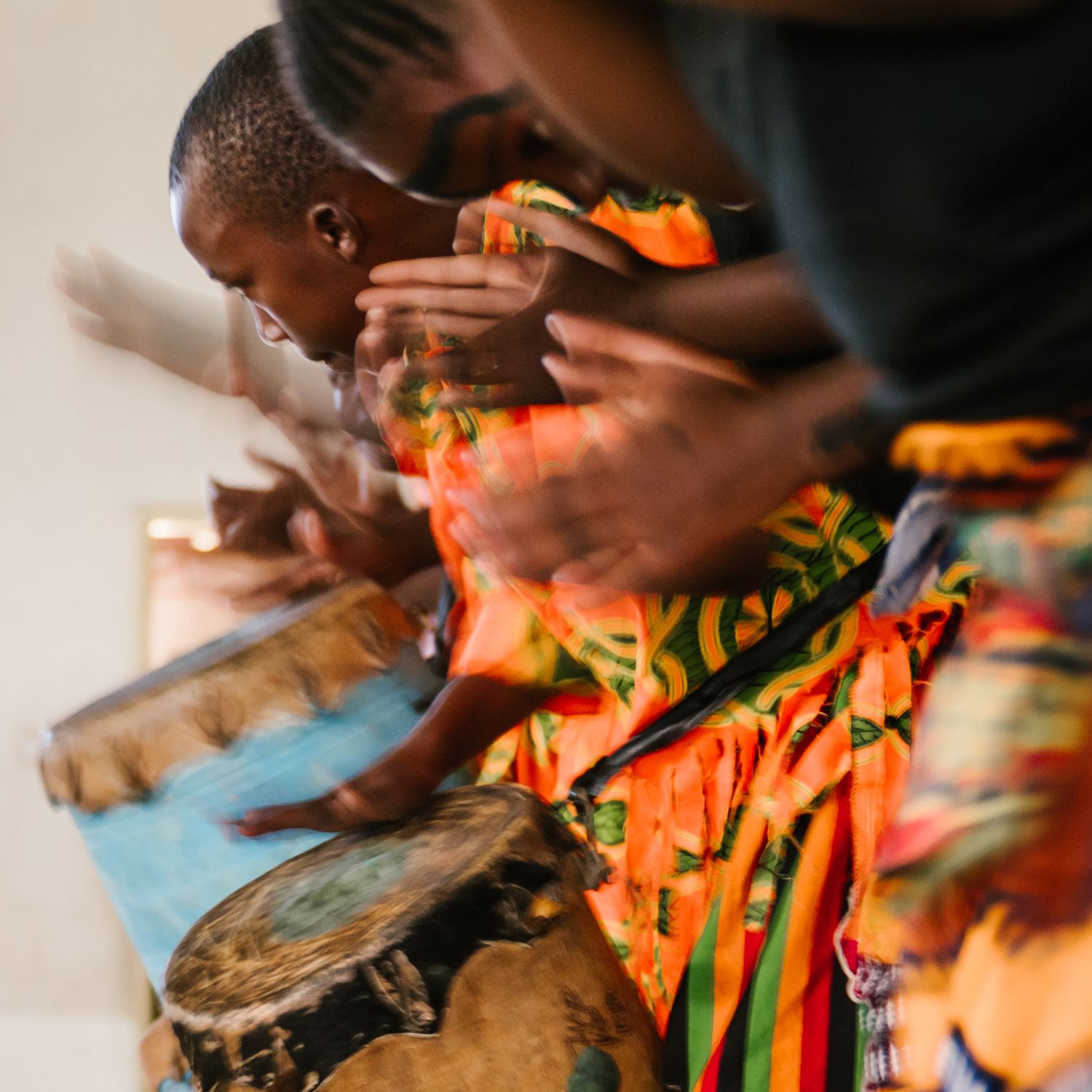 A musician in colorful kente cloth plays a djembe drum with dynamic motion blur during a traditional African performance.
