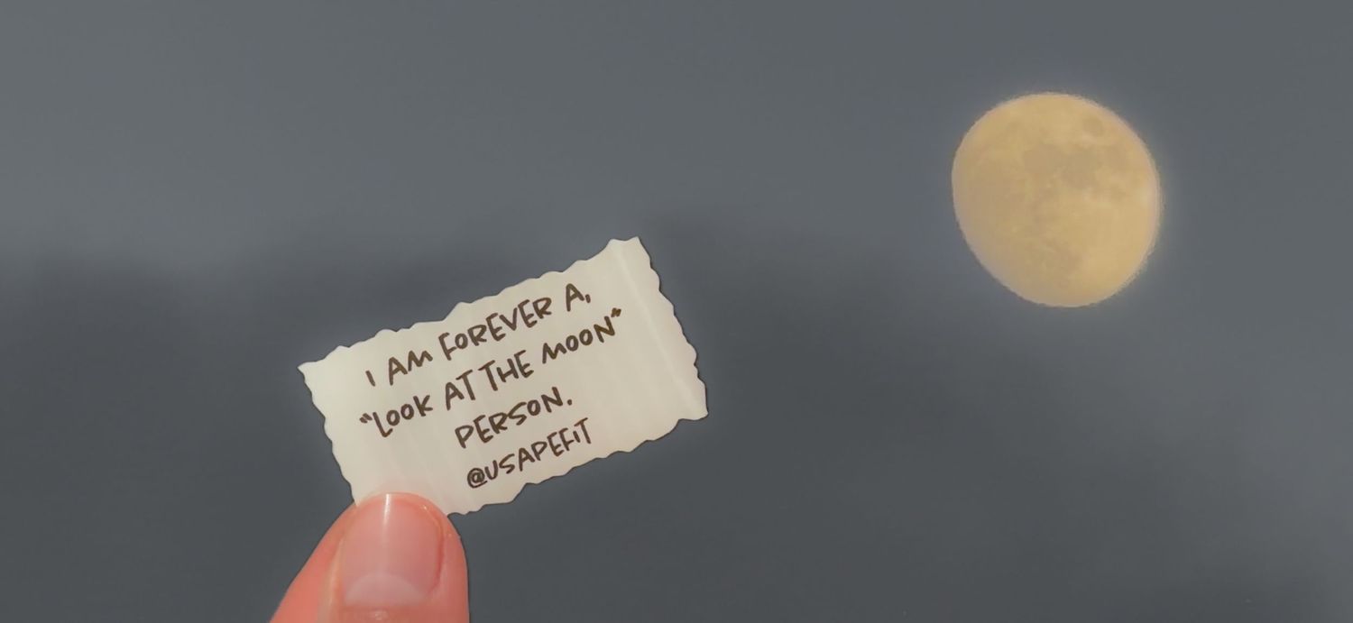 Hand holding a note saying 'I am forever a look at the moon person' with a full moon in cloudy sky.