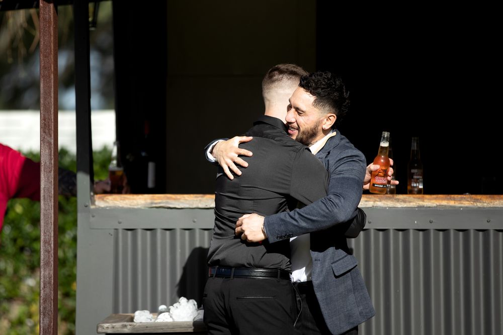 Two men in suits share a warm hug outdoors near a bar with bottles in the background.