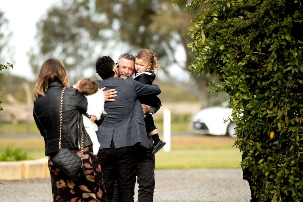 People in formal black attire sharing warm embraces outdoors at a gathering, with green hedges and cars in the background.