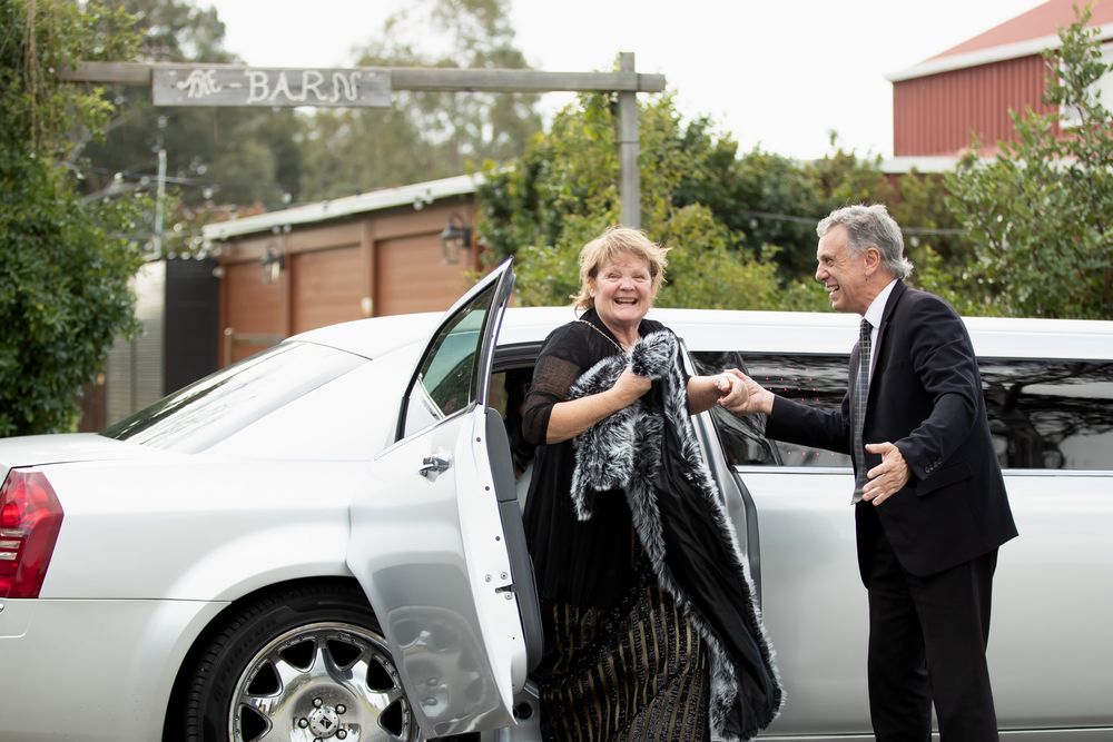 A woman in formal attire exits a white limousine as a suited man holds the door open outside a barn.