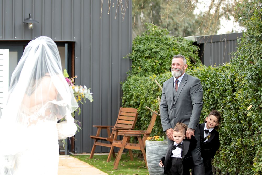 Groom smiling as bride approaches in white gown and veil, with two young boys in suits at outdoor wedding ceremony.