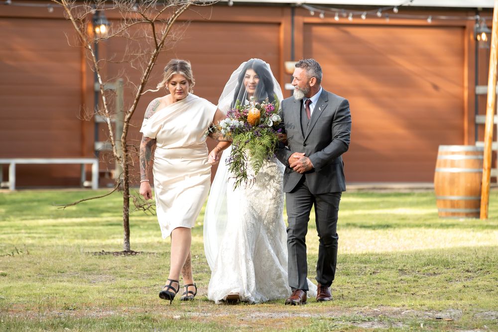 Bride in white lace gown and veil walks with parents carrying bouquet at outdoor wedding venue.