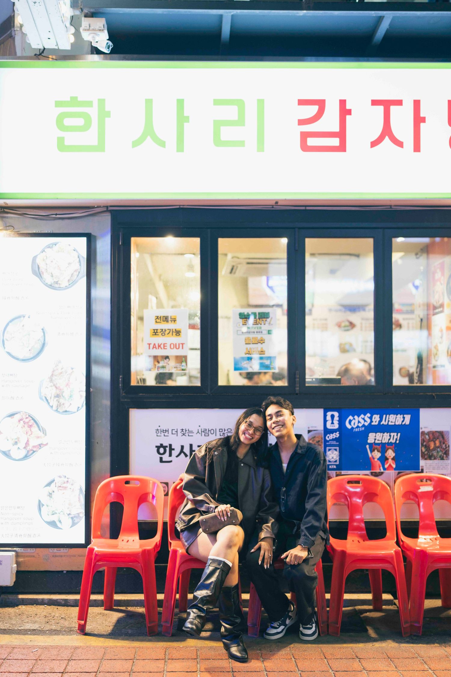 Couple sitting together on red chairs outside a Korean restaurant at night, smiling warmly.