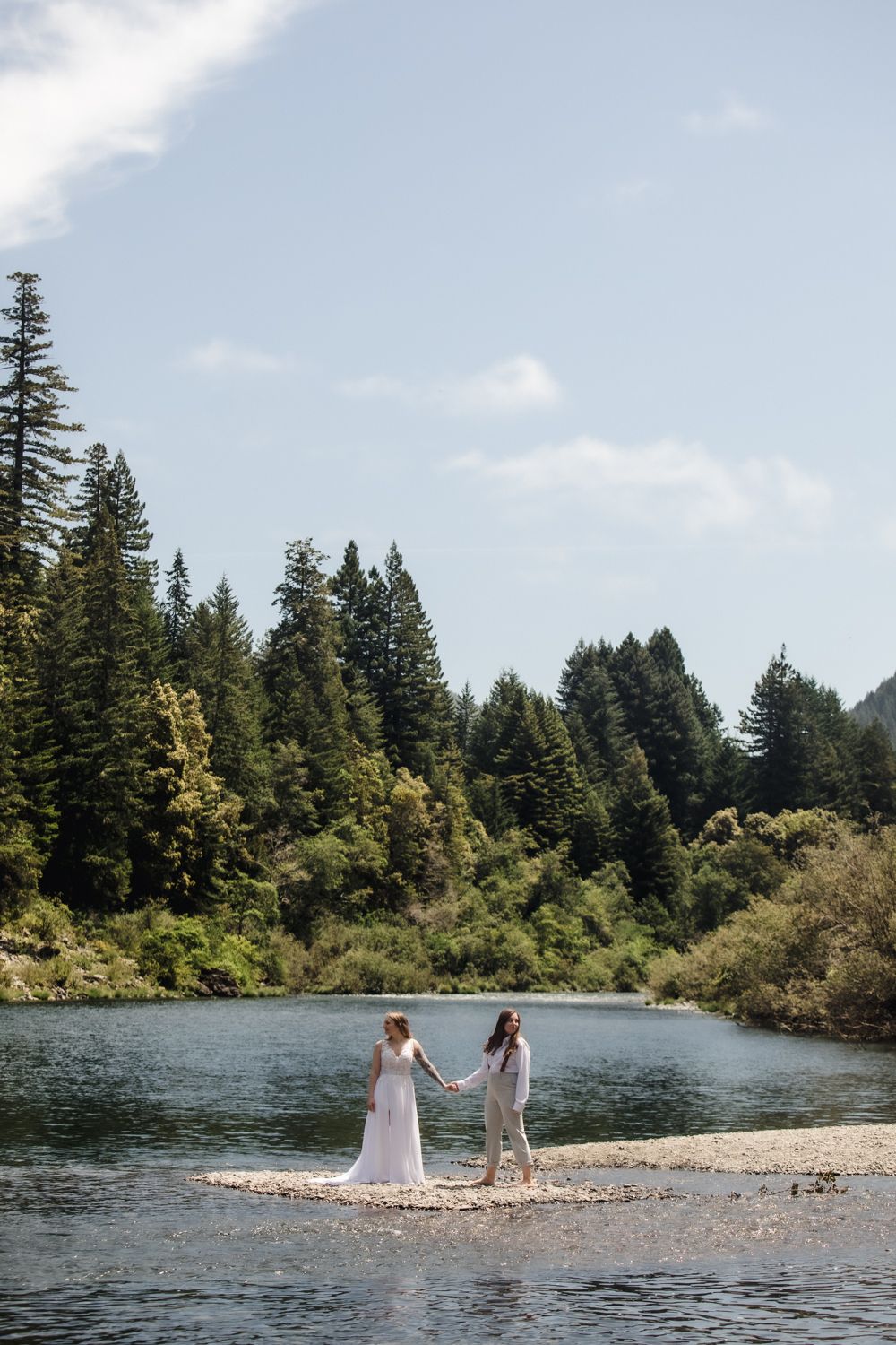 Wedding couple standing among towering redwood trees in a lush forest, bride in white gown and bride in suit.