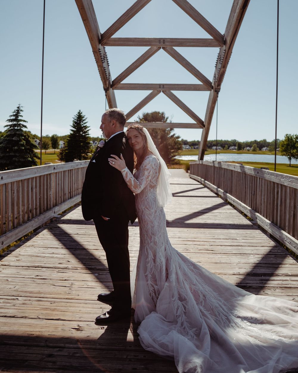 Bride and groom share a romantic moment on a wooden bridge, her lace gown trailing elegantly behind them.