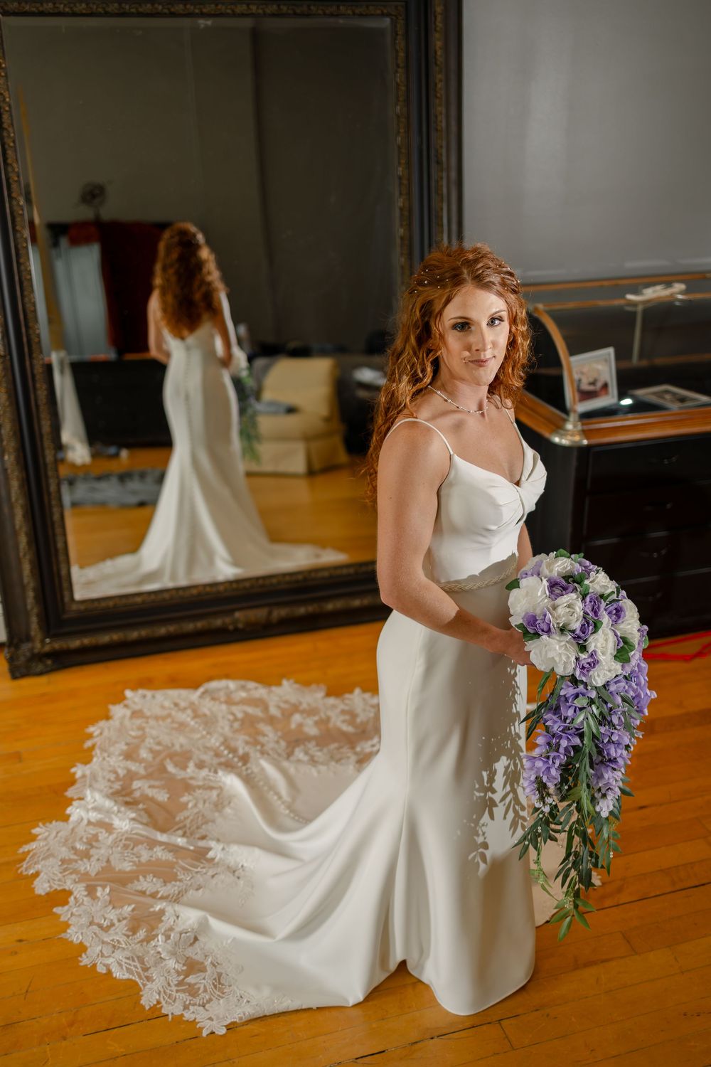 Redheaded bride in a white satin gown with lace train holds purple floral bouquet, reflected in ornate mirror.