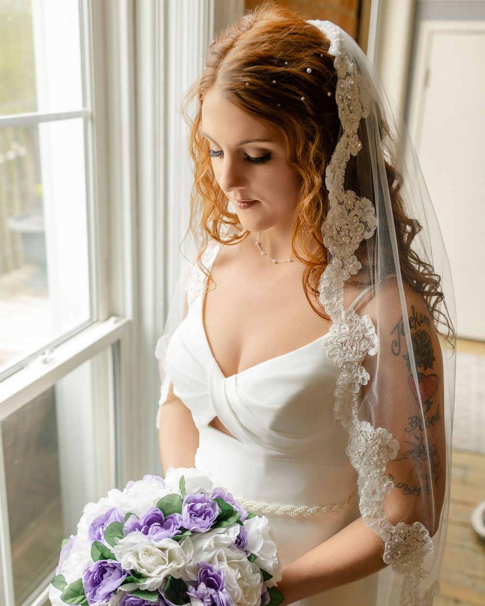 Bride in white gown and lace veil holds purple and white bouquet by window on wedding day.