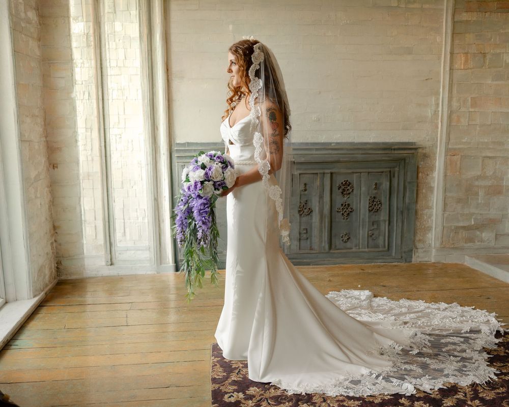 Bride in white gown with lace veil holds purple bouquet, standing by tall window in elegant room with hardwood floors.