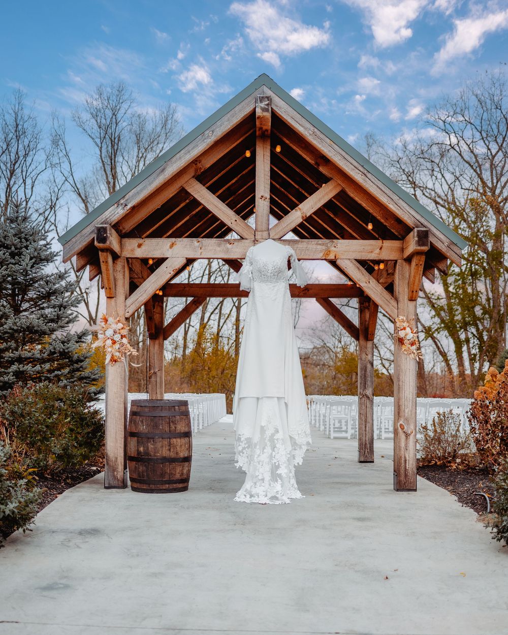 A lace wedding dress hangs under a rustic wooden pavilion with floral arrangements and a barrel, set against a blue sky.