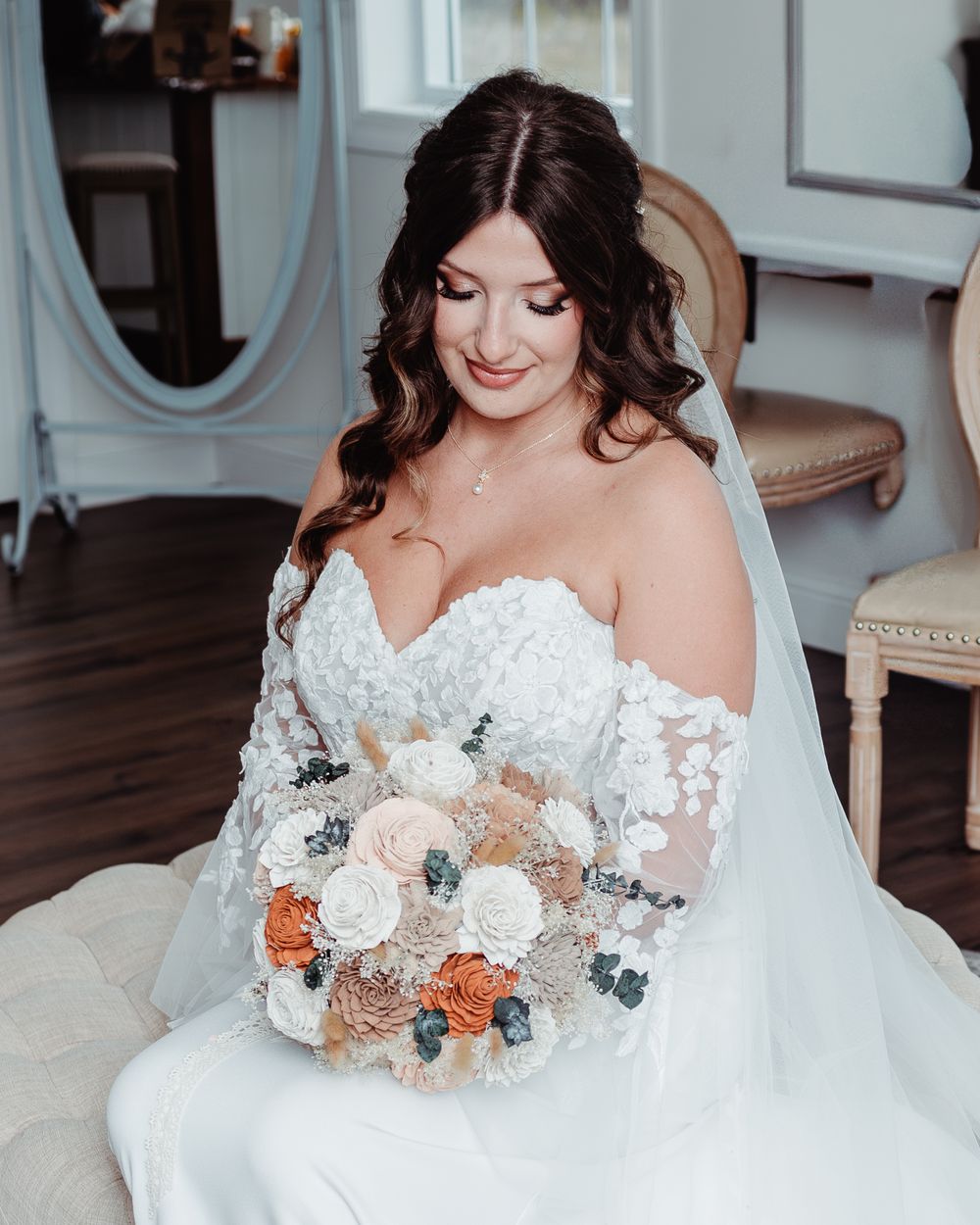 Bride in off-shoulder lace gown holds bouquet of white, blush, and orange flowers while sitting and smiling softly.