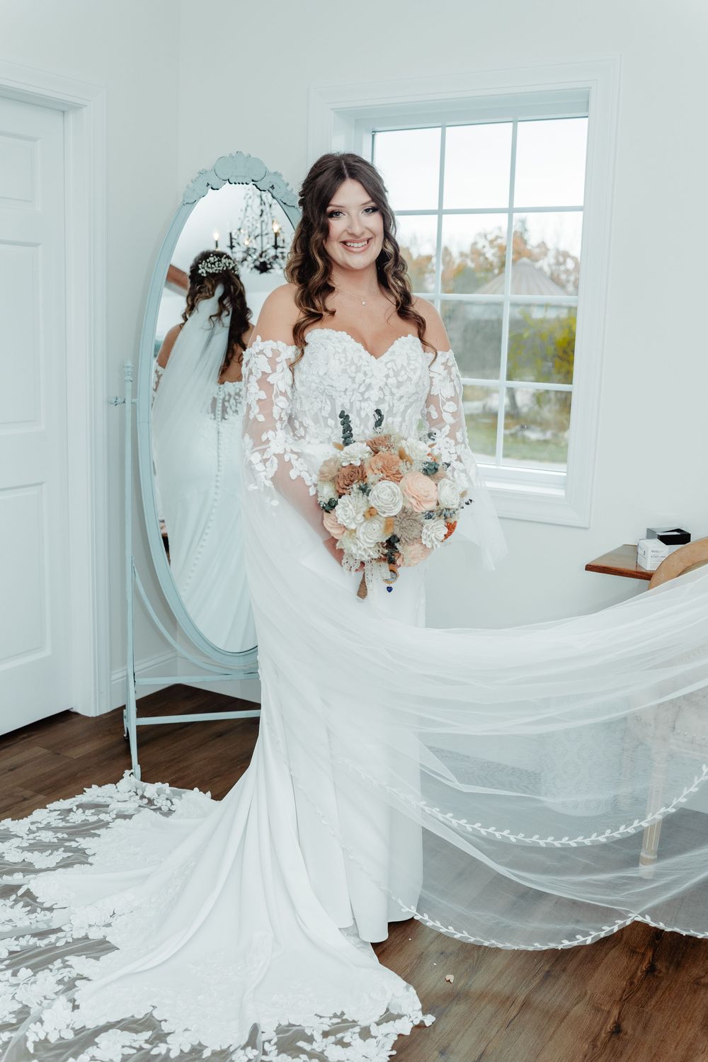 Bride in lace off-shoulder wedding gown holds bouquet before ornate mirror in bright white bridal suite.