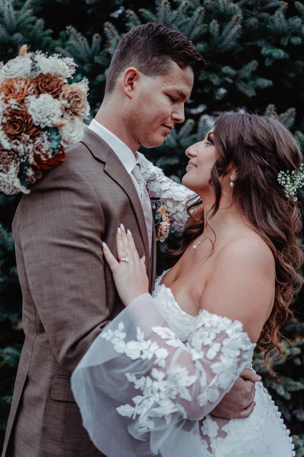 Bride and groom share romantic moment embracing outdoors in front of evergreen trees on wedding day.