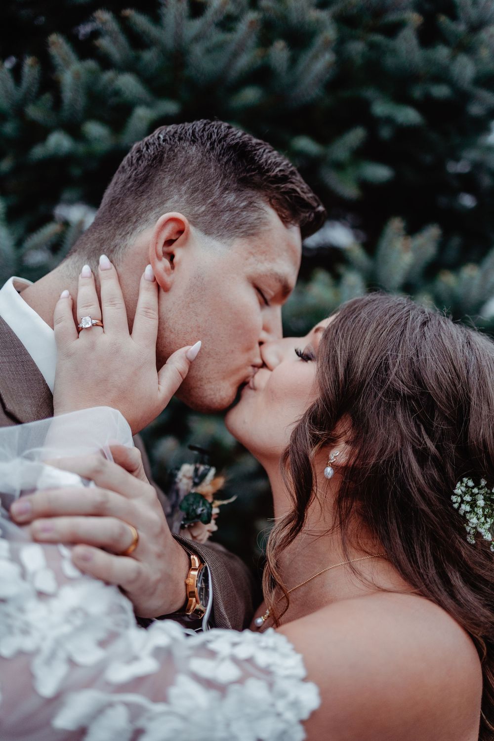 A bride and groom share a romantic first kiss at their wedding ceremony with evergreen trees in the background.