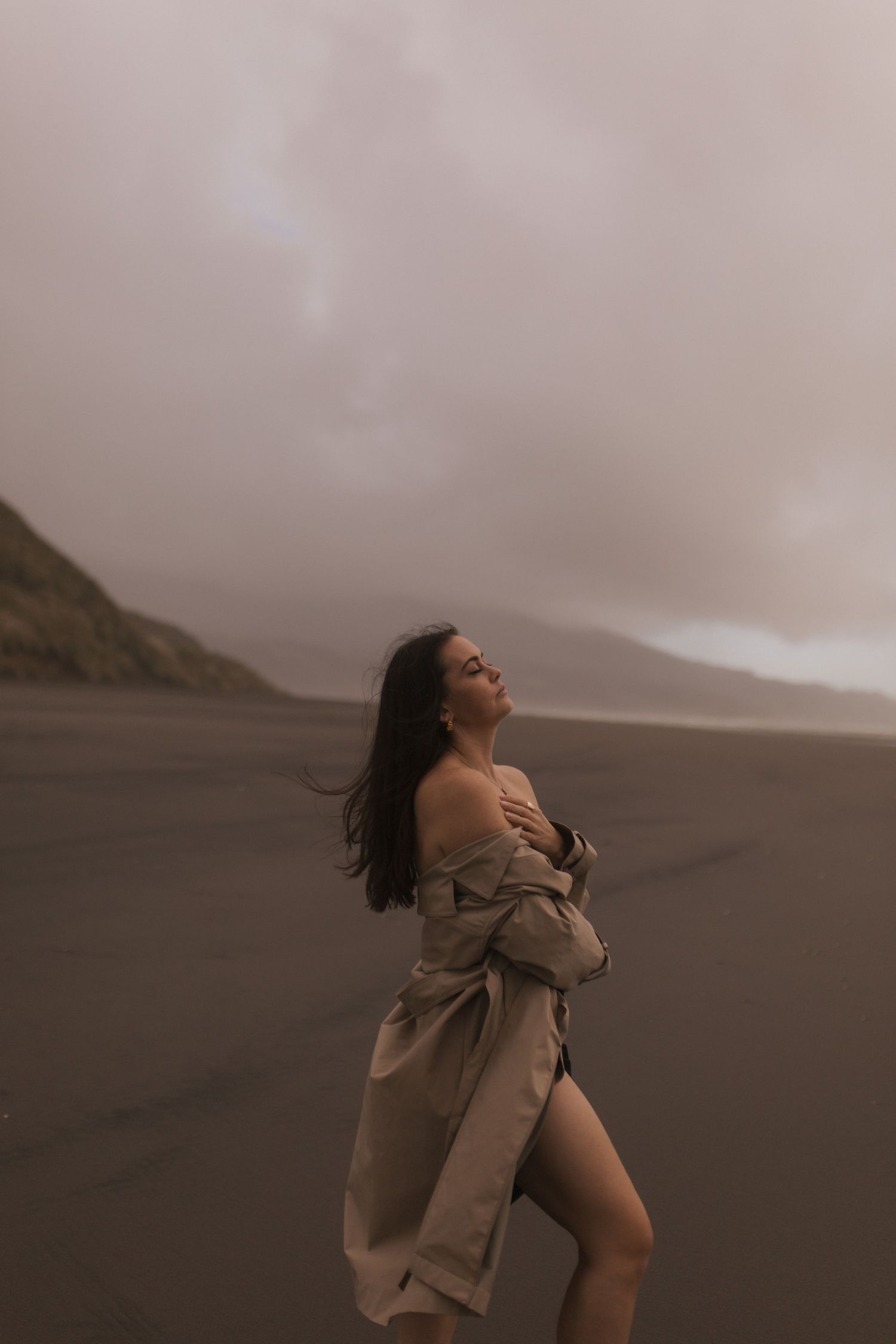 Woman in beige coat on dark sand beach with misty mountains, hair blowing in wind, moody overcast sky.