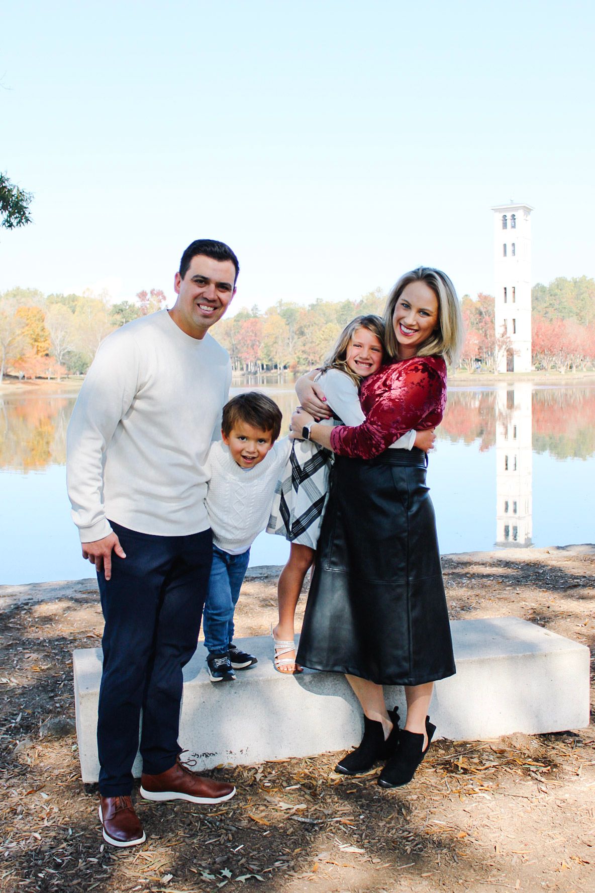 Happy family of four posing outdoors by a serene lake with autumn foliage and a tower reflection.
