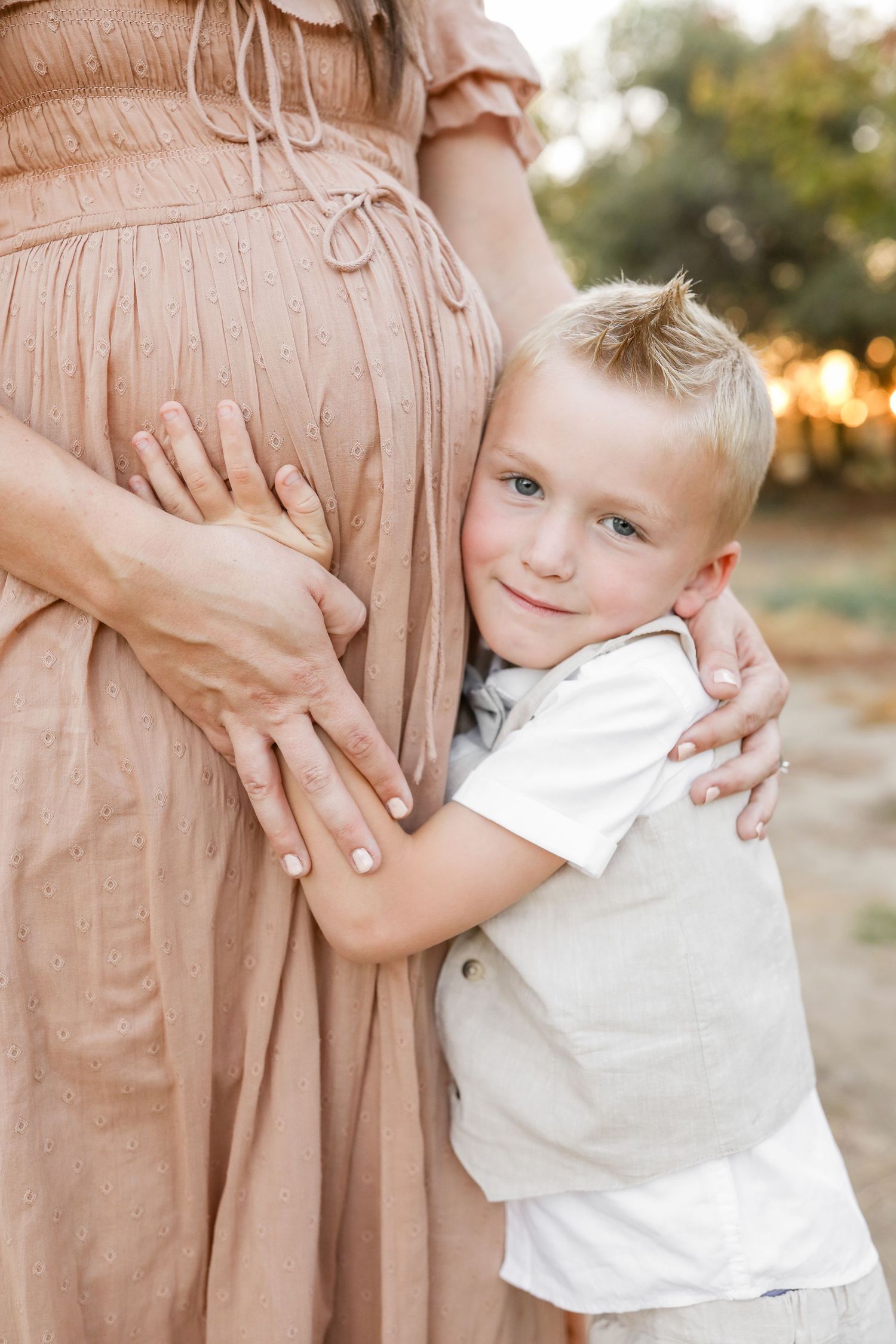 A young blonde boy hugs his pregnant mother's belly outdoors, wearing a white shirt and vest.