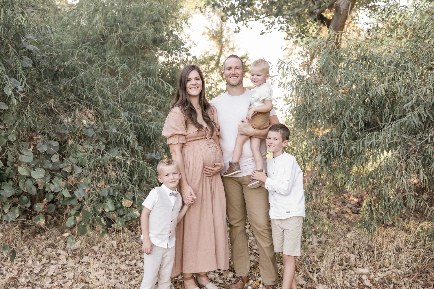 Pregnant woman in pink dress poses with husband and three children outdoors in a lush green nature setting.