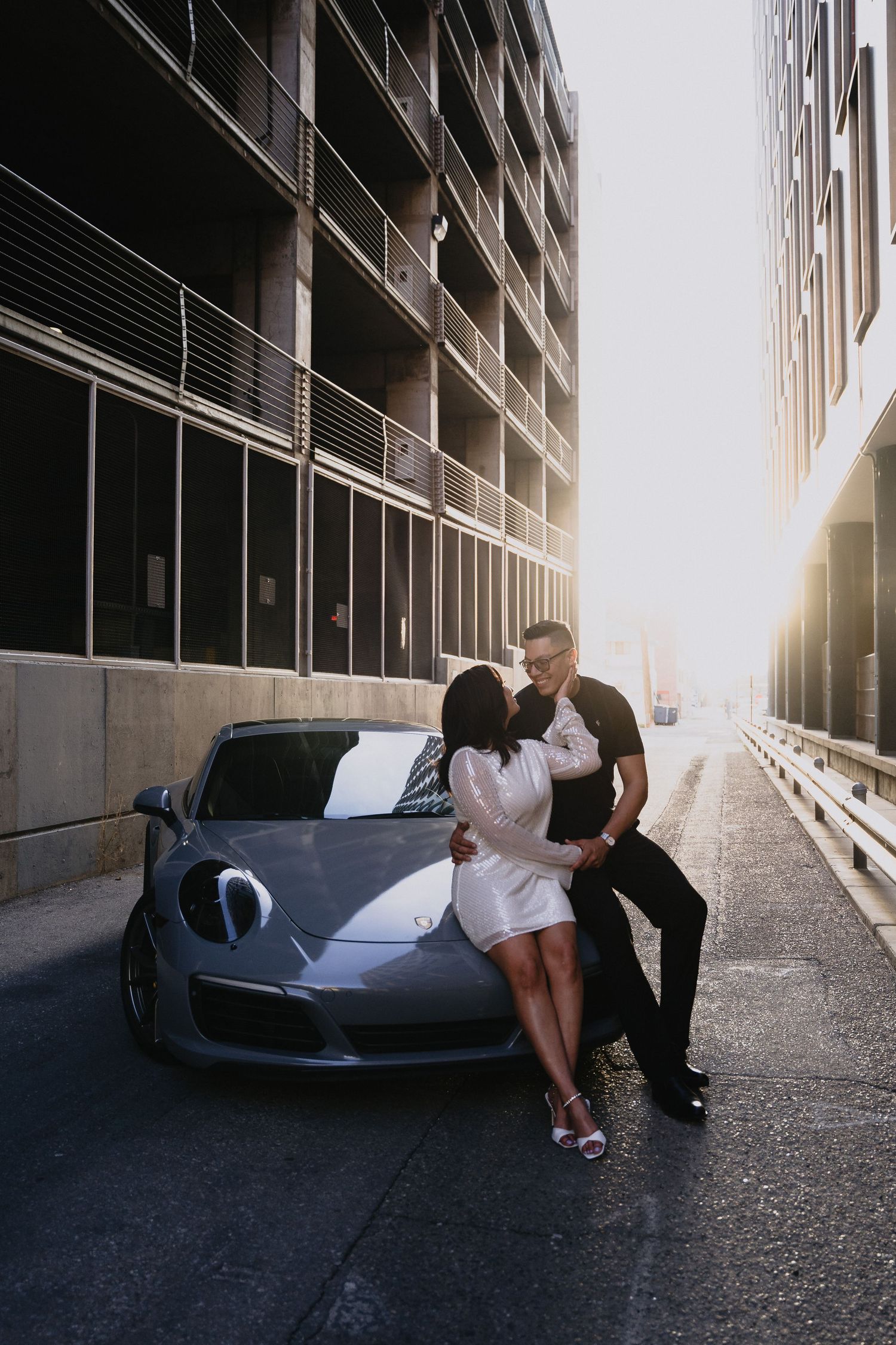 Couple embracing romantically against a sleek gray Porsche in an urban alley with golden sunset light in Downtown Albuquerque