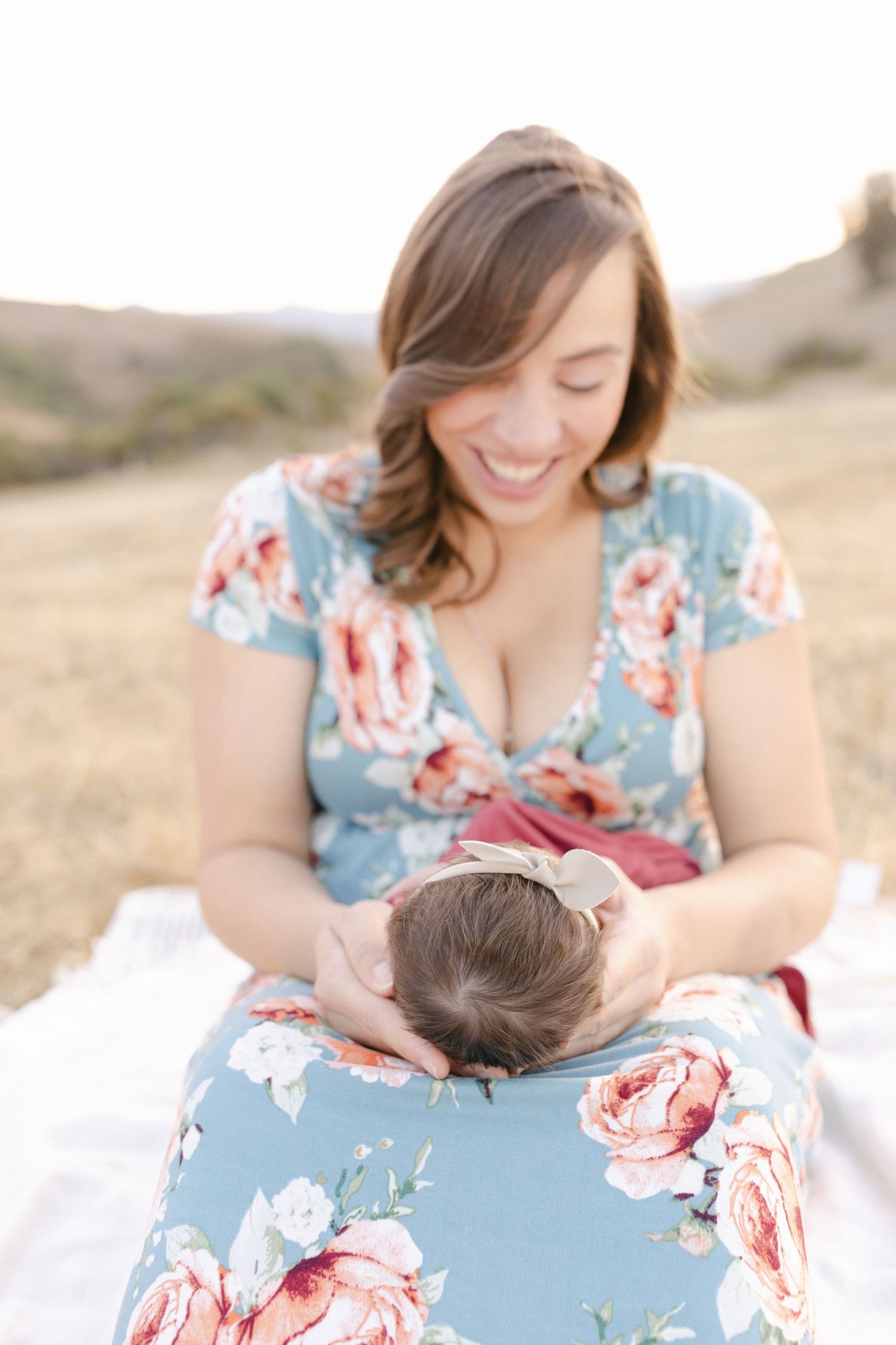 Smiling mother in floral blue dress cradling newborn baby outdoors in a sunlit field.