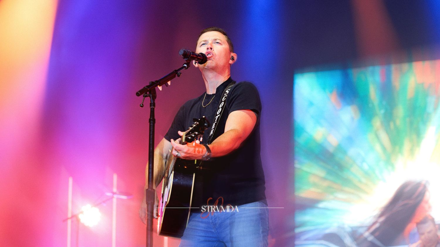 Male singer performs on stage with guitar at colorful concert, singing into microphone under vibrant light display.